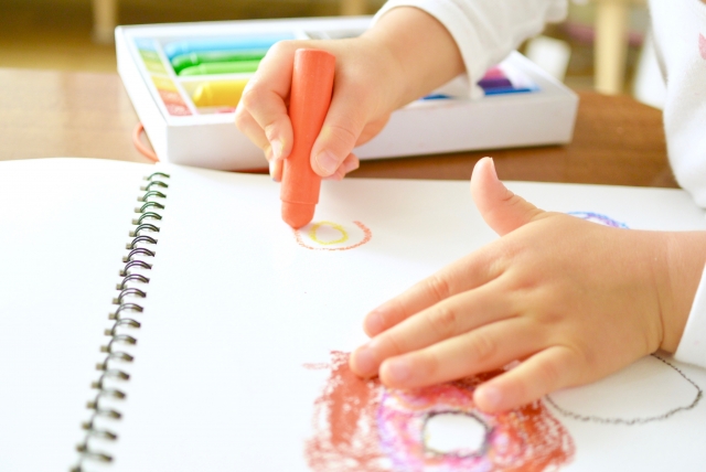 机の上のスケッチブックにクレヨンで丸を描く子どもの手元。