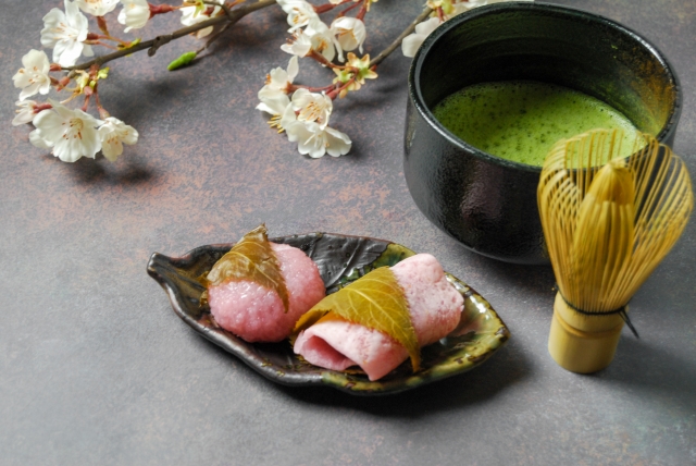 桜の花を背景に、桜餅と抹茶、茶筅を添えた春らしい和の一景